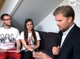 En mand, der holder en mikrofon, ser ud til at interviewe en siddende mand og kvinde. De siddende personer er iført matchende hvide T-shirts med grafik. Rummet har skråloft og et lille vindue. TV2 Fyns logo er synligt i hjørnet.
