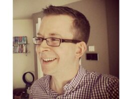 En person med kort hår og briller smiler, iført en ternet skjorte, i et værelse med en hylde i baggrunden.