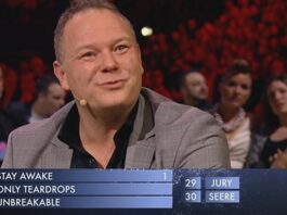 En mand i et gråt jakkesæt sidder ved et bord under en tv-begivenhed og smiler. Bag ham er et stort publikum synligt. En resultattavle viser sangtitler og partiturer: Stay Awake, Only Teardrops, Unbreakable, med forskellige point fra Jury og Seere.